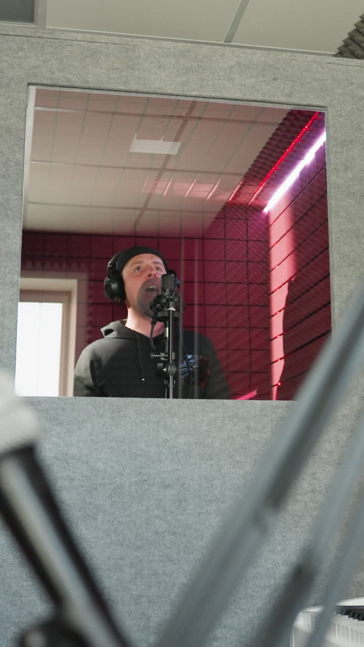 A man dressed in black stands in front of a studio microphone, wearing headphones as he sings passionately in a red soundproofed room. a recording session in a professional studio environment