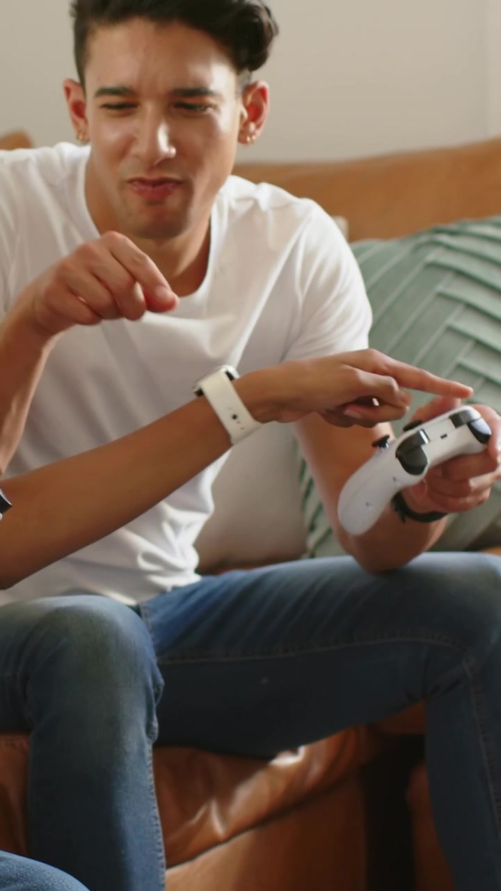 Vertical video: Leaning man guiding game controls while woman learning on sofa, white controller
