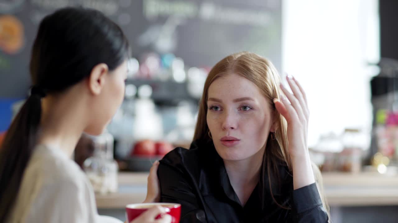 카페에서 커피를 마시며 이야기하는 두 젊은 여성의 오른쪽 을 추적합니다. 빨간 머리 백인 여성이 그녀의 문제에 대한 슬픈 이야기를 들려주고, 아시아 친구가 그녀를 위로하기 위해 어깨를 습니다.