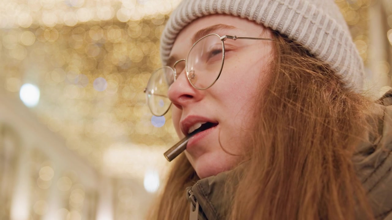 Donna che mastica una penna sotto le luci, occhiali rotondi e cappello di lana che incorniciano un'espressione concentrata mentre scrive testi in un mercato invernale, il primo piano cattura un'atmosfera pensierosa, un processo creativo e una luce soffusa.
