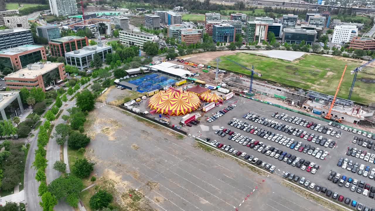 Orbiting drone aerial of colorful circus tent with carnival setup, large parking lot with cars, and urban commercial district backdrop