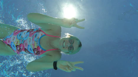 cheeky female athlete swimmer dives down to underwater camera, smiles and gives peace sign, 4k slow motion