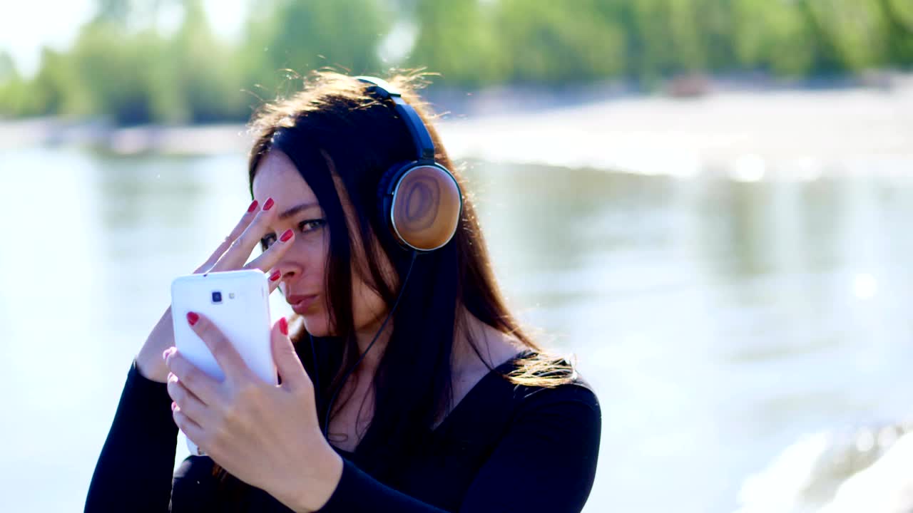 лето, возле реки, на пляже. на рассвете, красивая женщина, с длинными темными волосами, слушает музыку на наушниках с мобильного телефона, устройства, снимает себя на телефоне, делает селфи