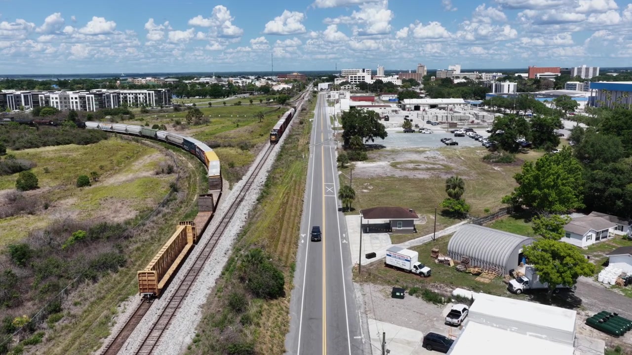Un treno merci curva lungo la ferrovia accanto a una strada dritta vicino a Lakeland, Florida, USA, incorniciato da magazzini, appartamenti, uno skyline del centro città in lontananza sotto un cielo nuvoloso e azzurro. Ripresa aerea con drone in sorvolo