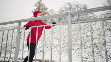 woman exercising outdoors during winter, stretching her leg back and forth with hand, wearing black gloves, background includes snowy ground, foggy atmosphere, and distant trees