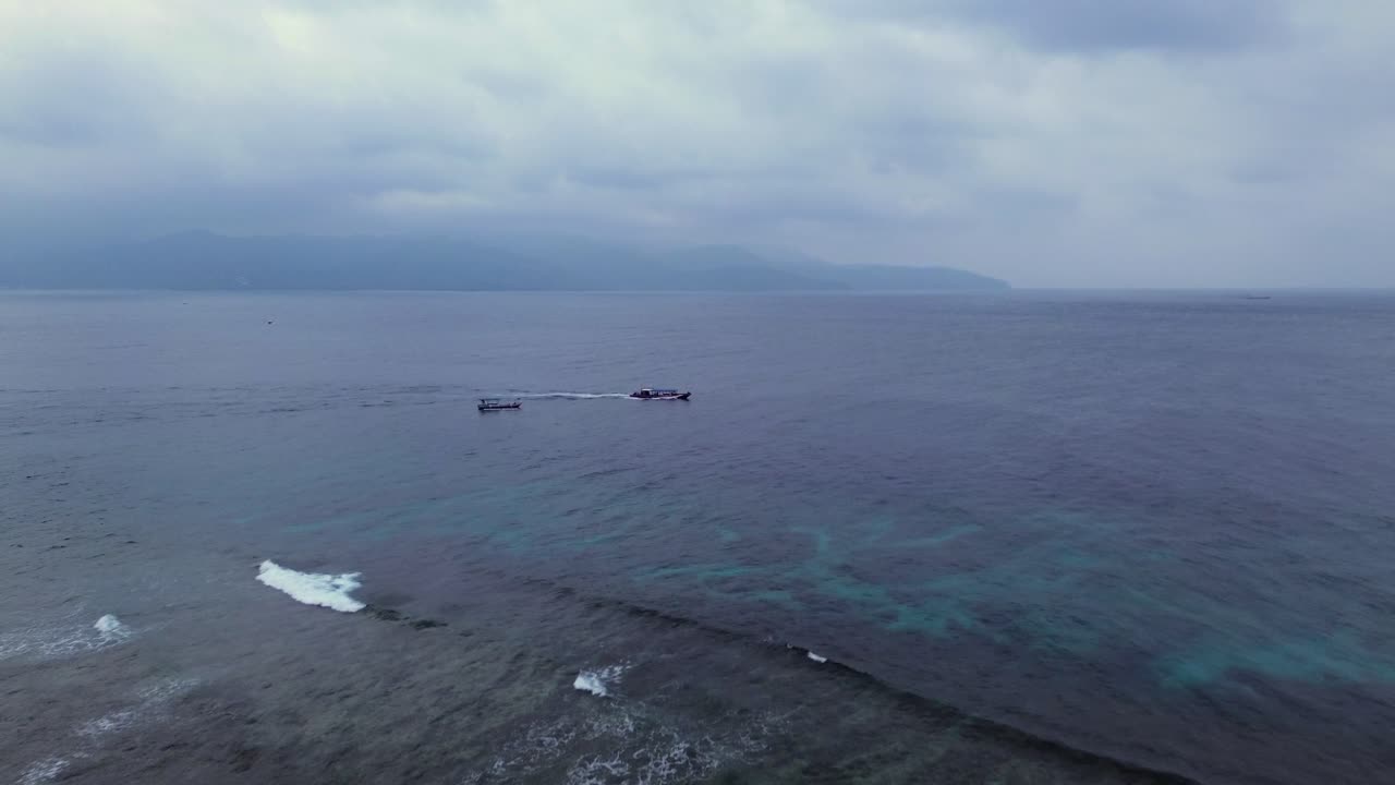 Aerial zoom in shot of boat cruising on calm ocean waters under cloudy skies near the Gili Islands, Indonesia