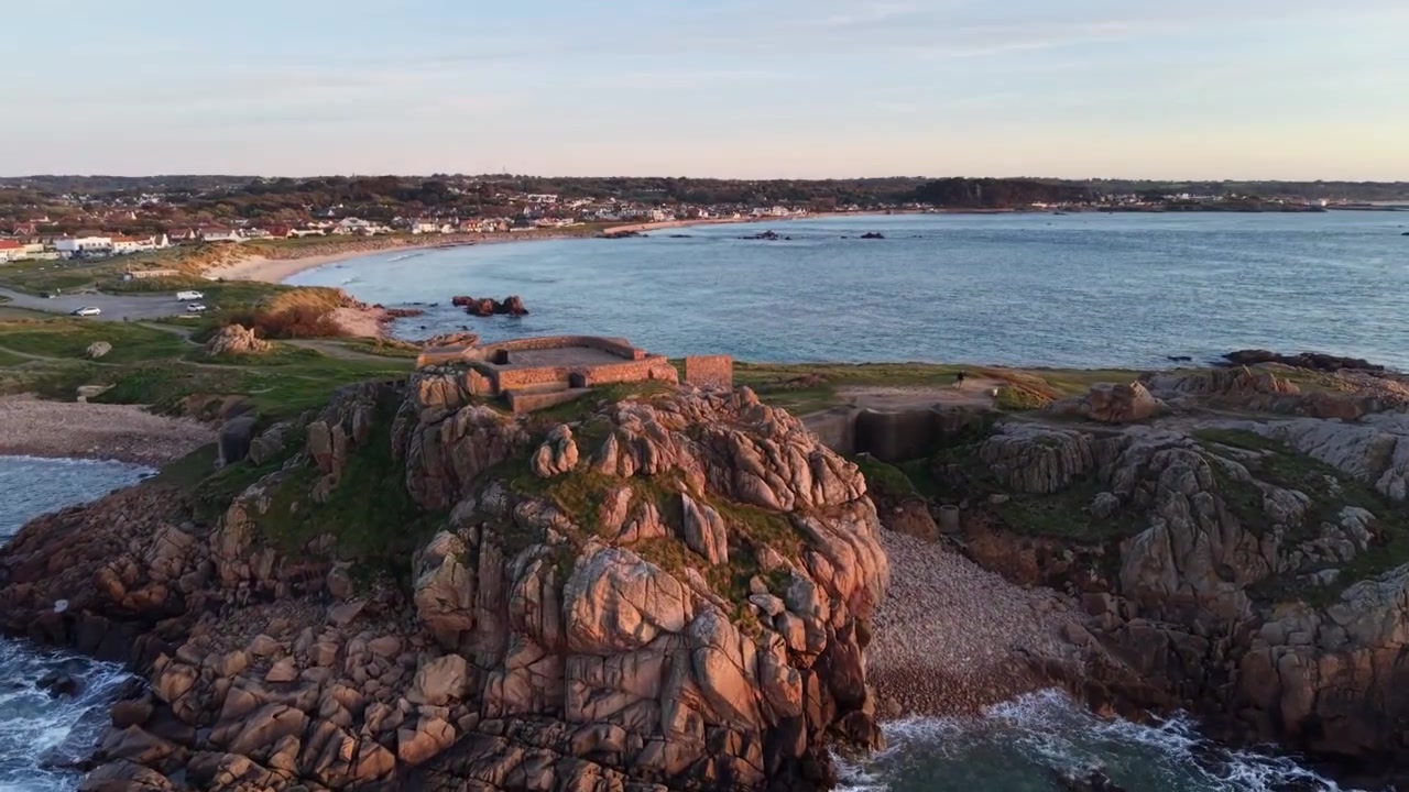 Grandes Rocques, Guernesey, Îles Anglo-Normandes. Images de drone à l'heure dorée s'élevant au-dessus d'une eau bleue claire, de rochers avec des vagues déferlantes, vers un ancien fort militaire et de magnifiques baies et plages sur la côte ouest