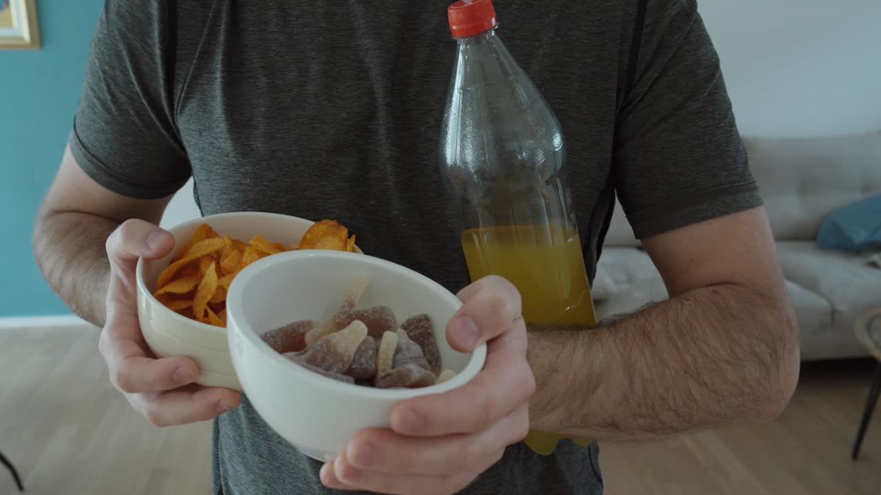 Uomo che tiene ciotole di patatine e caramelle insieme a una bottiglia di bibita zuccherata, illustrando scelte alimentari sbagliate, elevato apporto calorico e abitudini alimentari malsane a casa.