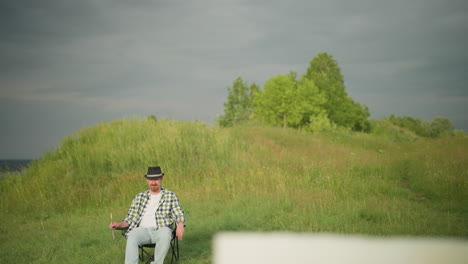 a man wearing a black hat, checkered shirt, and jeans sits comfortably on a chair in a sunlit, grassy field. the serene environment and casual attire suggest a peaceful moment in nature