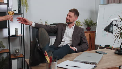 businessman drinking champagne from glass after working, putting legs on table sitting at office