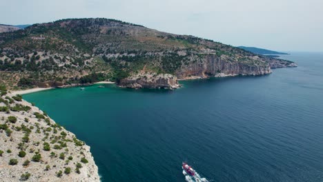 Aerial-View-Over-A-Seaside-Cliff-With-Turquoise-Water-And-Small-Beaches-With-The-Monastery-of-the-Archangel-Michael-In-The-Background,-Red-Boat,-Thassos,-Greece
