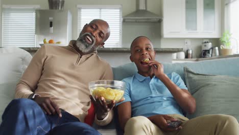 Video-of-happy-african-american-father-and-son-eating-chips-and-watching-tv