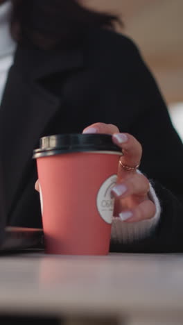 primer plano de la mano de la mujer sosteniendo suavemente una taza de café roja mientras escribe en la computadora portátil, mostrando una manicura elegante y un espacio de trabajo moderno con un fondo borroso de una cafetería o oficina al aire libre