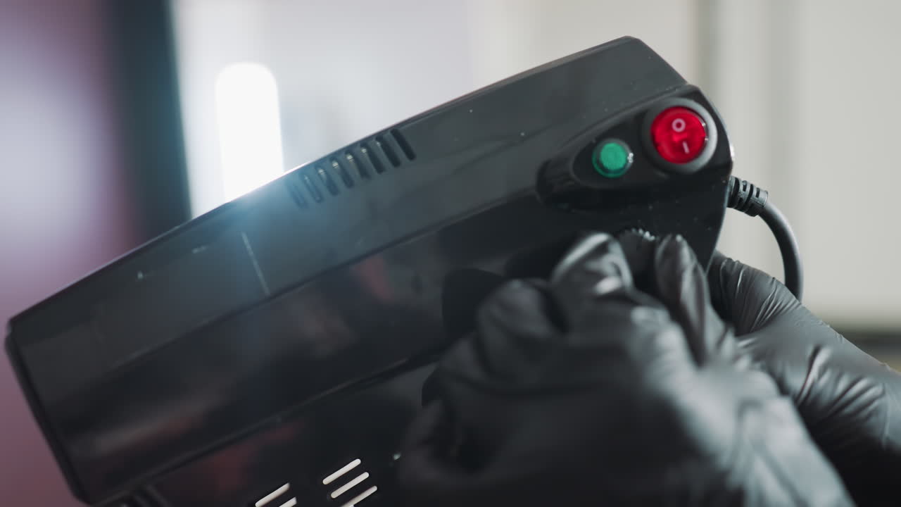 Mano enguantada ajustando una pistola de esterilización, entorno clínico con indicadores rojos y verdes, guante protector negro, primer plano de los botones de control y el cable, procedimiento y equipo de higiene precisos.