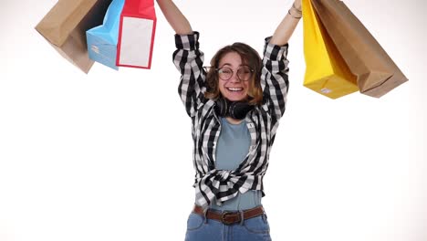 cheerful beautiful european young woman in glasses and headphones on neck holding colorful shopping bags in season sale, happily shakes them isolated on white background