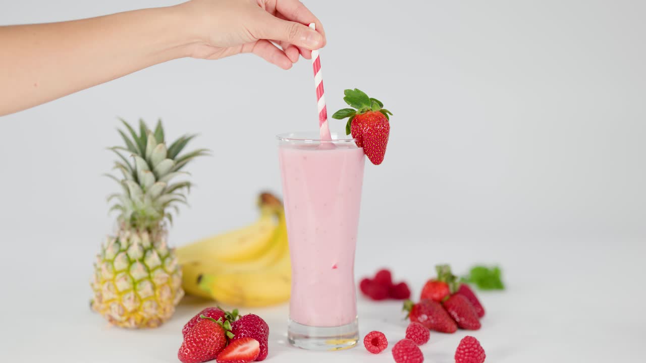 Hand stirs creamy pink smoothie in glass, surrounded by fresh strawberries, bananas, pineapple, raspberries