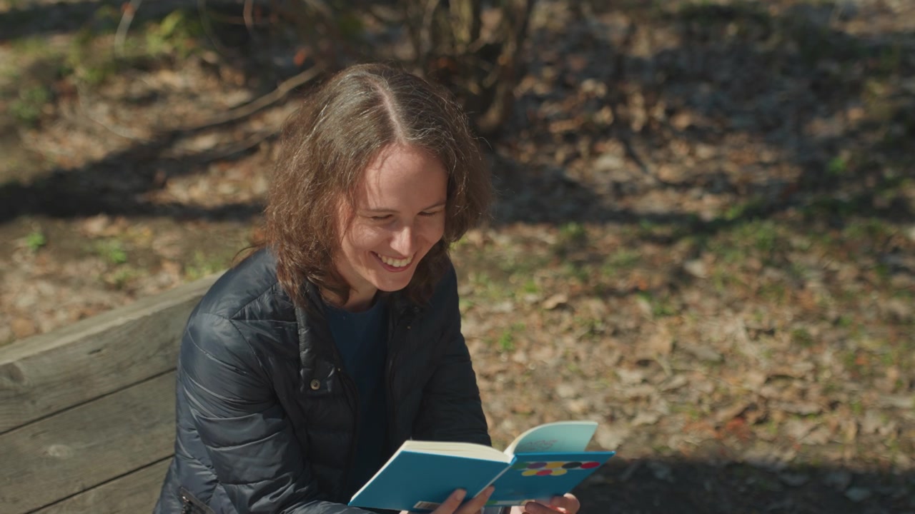Primo piano di una donna che legge un libro aperto mentre è seduta su una panchina di legno alla luce del giorno. La donna indossa una giacca scura e tiene un quaderno con copertina blu e cerchi colorati che mostra coinvolgimento attraverso il suo