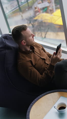 businessman relaxing on view from office window. guy with a phone looking at city through window on comfortable ottoman. work life balance concept. top view. vertical video