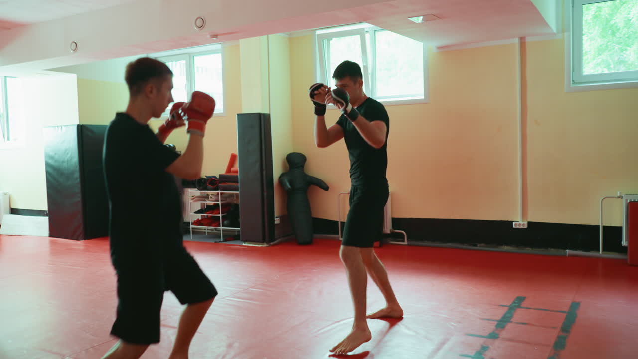 Two male fighters training martial arts in gym fighting face to face wearing boxing gloves preparing for sparring session on red mat floor under natural window light with equipment in background