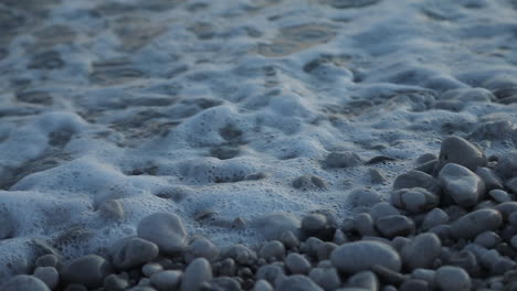 Foam-over-pebbles-on-seashore,-beautiful-texture-with-salty-water-moving-slowly