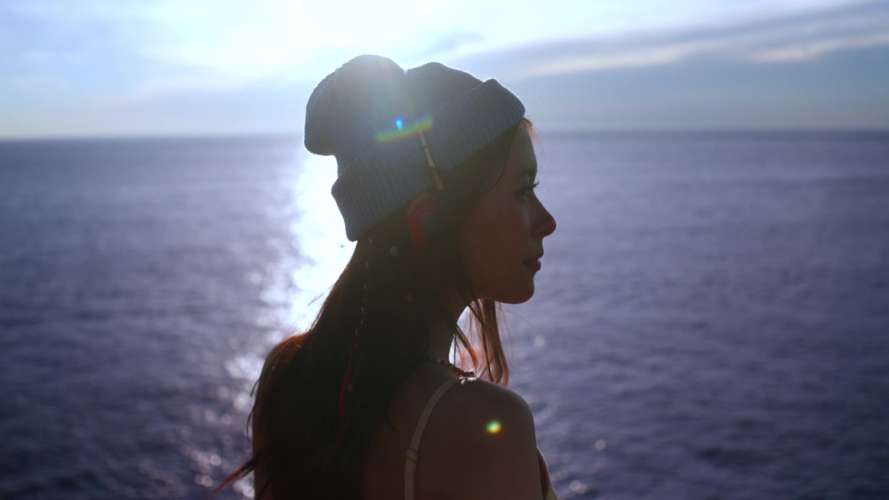 Young woman in a blue beanie stands in profile against a sunlit ocean horizon at sunset, beautiful longing facial expression with soft smile