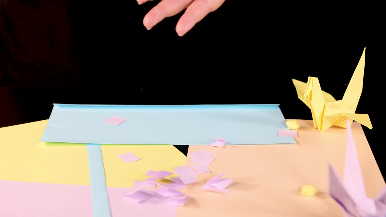 Hand drops small origami paper squares onto pastel paper surface with origami cranes, studio lighting