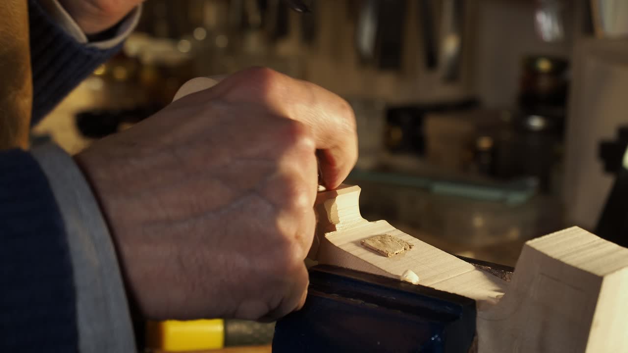 Extreme close up showing luthier’s hands carefully carving the volute of a violin scroll with a curved gouge chisel in his workshop