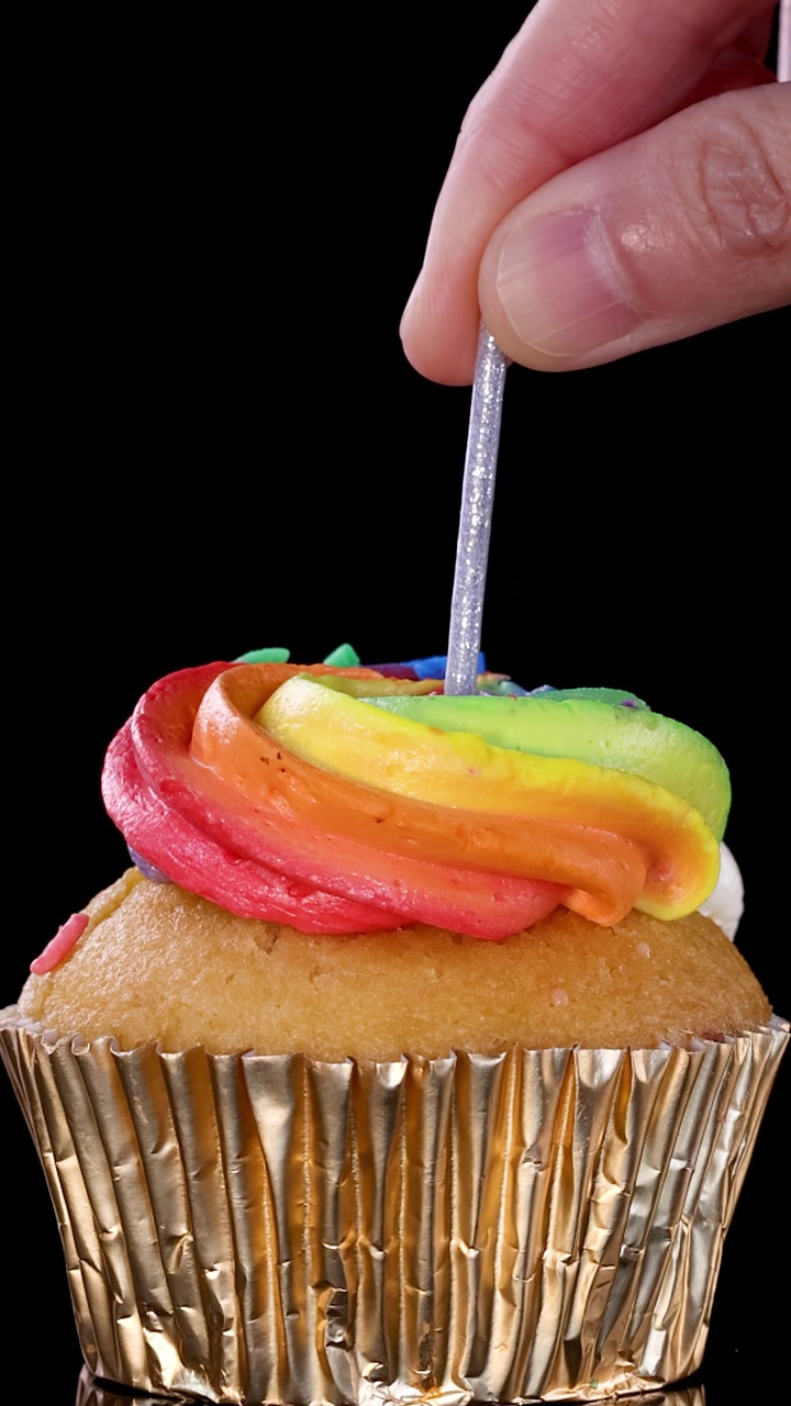 Una mano inserisce una candela argentata in un cupcake con glassa arcobaleno e pirottino dorato, su uno sfondo nero con illuminazione brillante e uniforme.