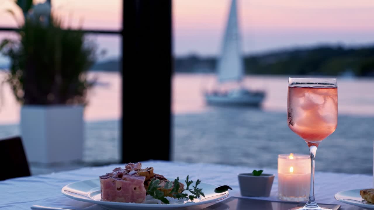 Romantic sunset dinner by the sea is served with a delicious dessert, a glass of rose wine, and a lit candle, creating a magical atmosphere with a sailboat in the background