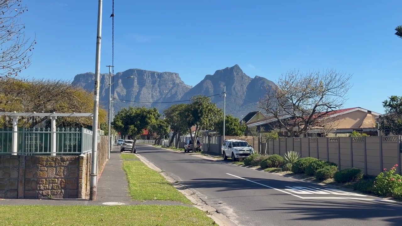Una tranquilla strada residenziale a Città del Capo, fiancheggiata da proprietà recintate, auto parcheggiate e alberi frondosi, con la Table Mountain e il Devil’s Peak che si ergono imponenti sullo sfondo sotto un cielo azzurro e limpido