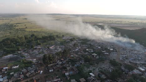 Captivating-4K-aerial-footage-features-a-push-in-drone-shot,-seamlessly-revealing-a-rural-township-village-situated-on-the-edge-of-a-waste-processing-plant-burning-waste-in-South-Africa