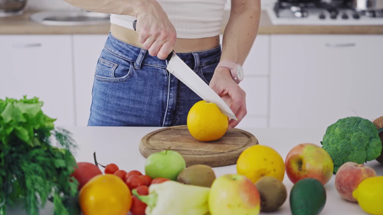 Woman with sharp knife cutting orange into two halves. Table full of fresh fruit and vegetables in the kitchen. Woman preparing healthy food for losing weight.