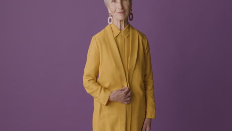 Mujer-Mayor-Con-El-Pelo-Corto-Con-Camisa-Y-Chaqueta-De-Color-Mostaza-Y-Pendientes-Posando-Y-Sonriendo-A-La-Cámara-Con-Fondo-Morado-1