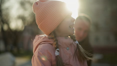 a close-up of a cheerful little girl in a pink cap and jacket smiling brightly with a man smiling in the background. the sunlight creates a warm, glowing effect