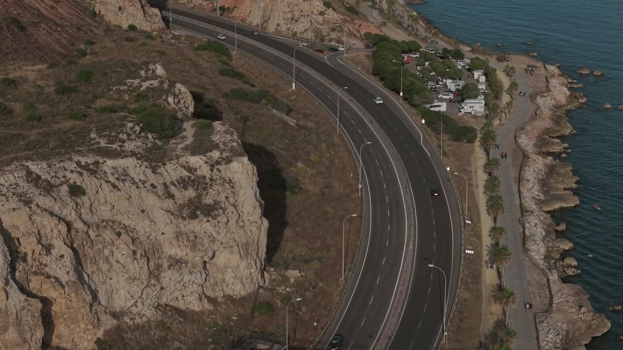 Coastal highway in Málaga curving along rocky cliffs by the Mediterranean Sea, with cars, a footbridge, and scenic oceanfront views at golden hour
