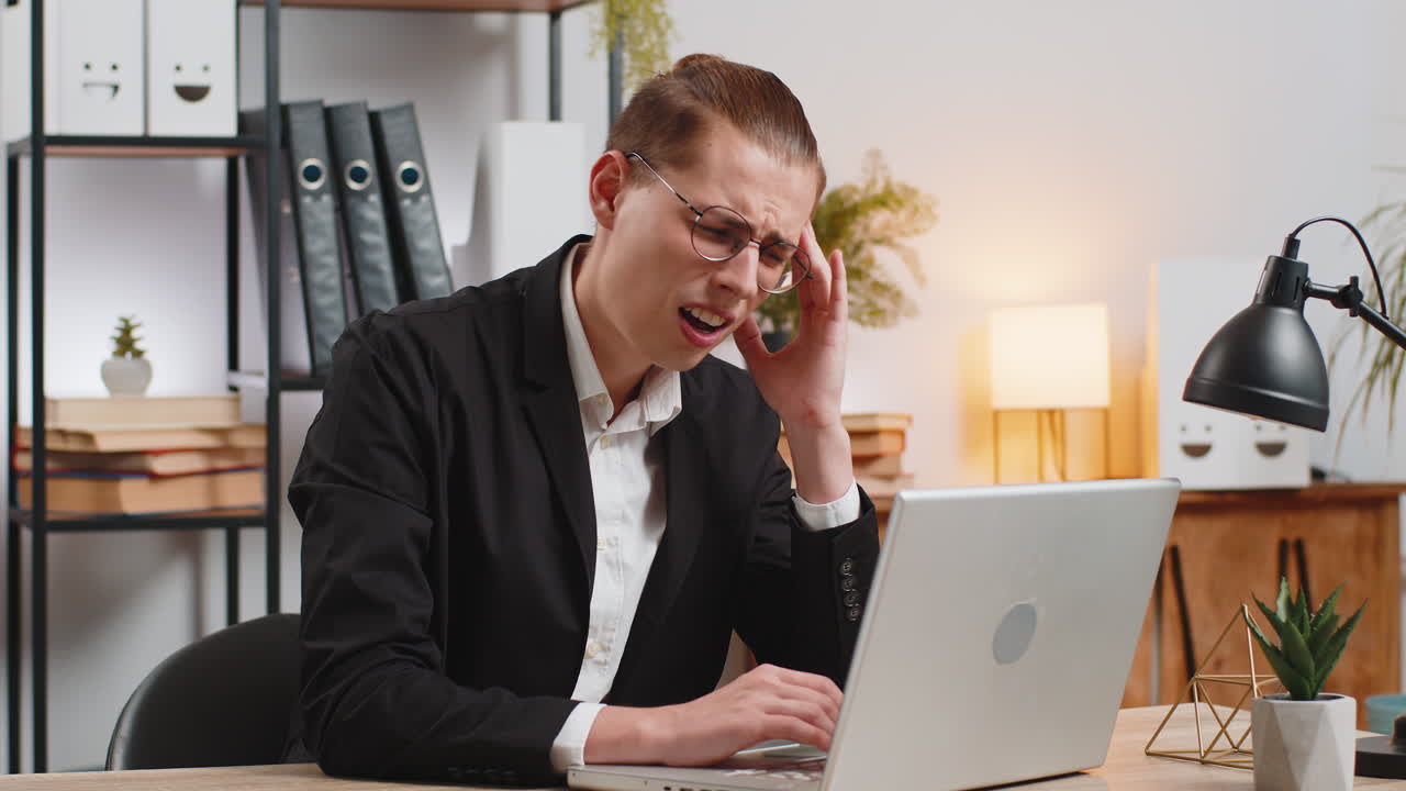 Sick young businessman working on laptop feeling headache rubs temples sitting at home office desk