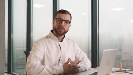 Gutaussehender-Und-Respektabler-Männlicher-Arzt-In-Einem-Weißen-Kittel,-Der-An-Einem-Schreibtisch-Mit-Einem-Laptop-In-Einer-Modernen-Klinik-Mit-Panoramafenstern-Sitzt-Und-Mit-Einem-Kunden-Spricht