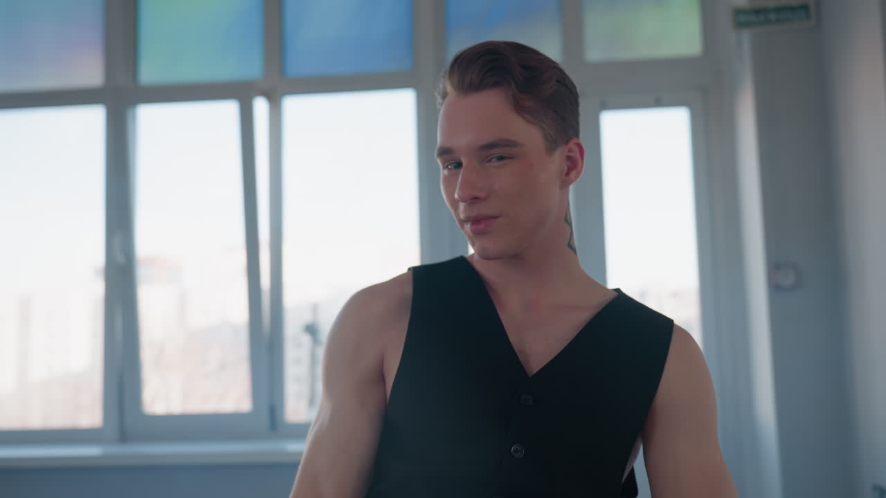 young man in black vest and top hat spins during break dance moves turning mid step to remove cap and wink at camera in loft studio with large windows and smoky mirror reflecting his silhouette
