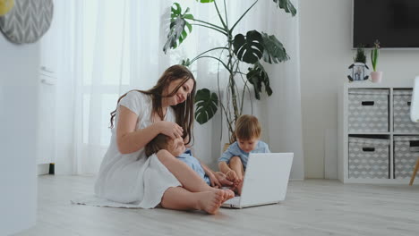 Una-Madre-Amante-De-Un-Apartamento-Moderno-Y-Dos-Hijos-Pequeños-Sentados-En-El-Suelo-De-La-Sala-De-Estar-Miran-La-Pantalla-Del-Portátil.-Los-Niños-Con-Mamá-Juegan-En-Una-Computadora-Portátil.