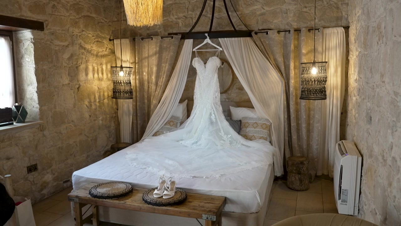 Un bellissimo abito da sposa è elegantemente appeso sopra un letto in una stanza rustica con pareti in pietra e illuminazione soffusa, creando un'atmosfera romantica perfetta per un giorno speciale.