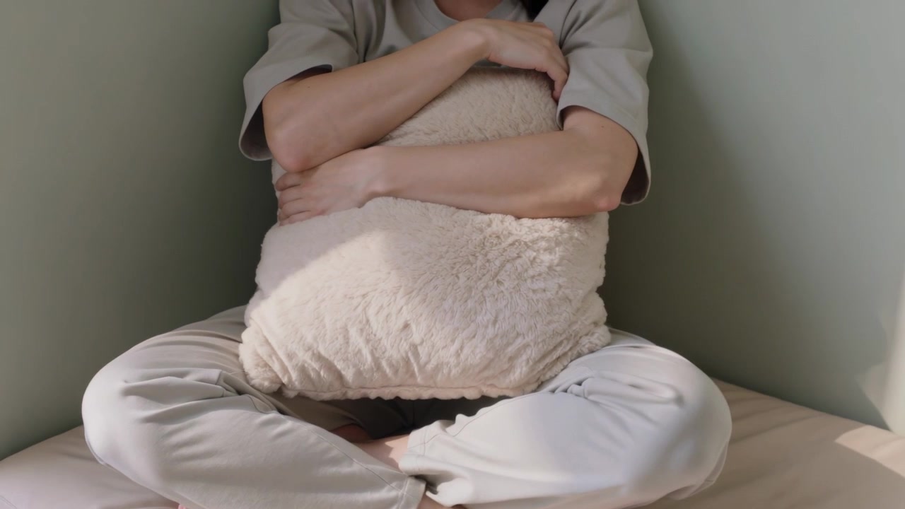 Hugging pillow, woman in beige outfit adjusting pose in bedroom corner settling as light brightens