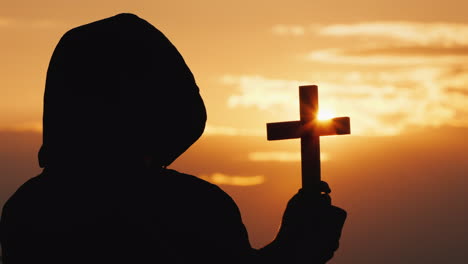 a monk in a hood with a crucifix in his hands stands against the backdrop of a dramatic sky at sunse