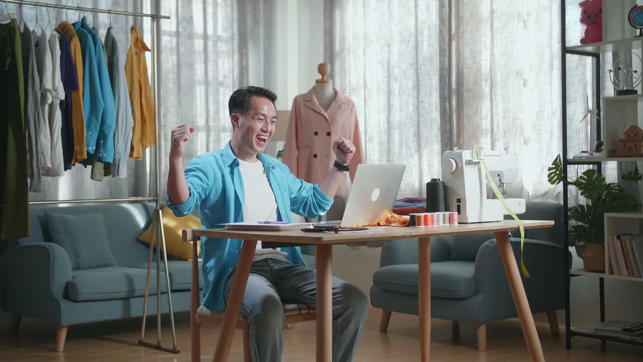 행복한 아시아 남성 디자이너가 노트북에서 일하고 스튜디오에서 옷을 디자인하는 동안 축하하고 있습니다.