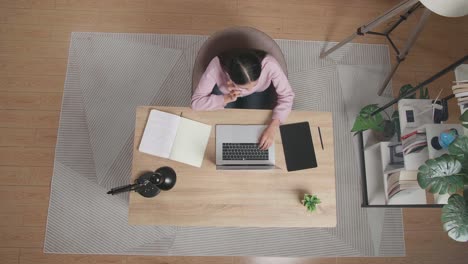 vista superior de una mujer hablando en un teléfono inteligente mientras está sentada en el escritorio de madera escribiendo en una computadora portátil en casa