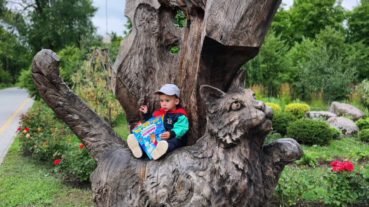 Sweet little boy sits on the wooden sculpture outdoors. Toddler touches the wood and looks at his book.