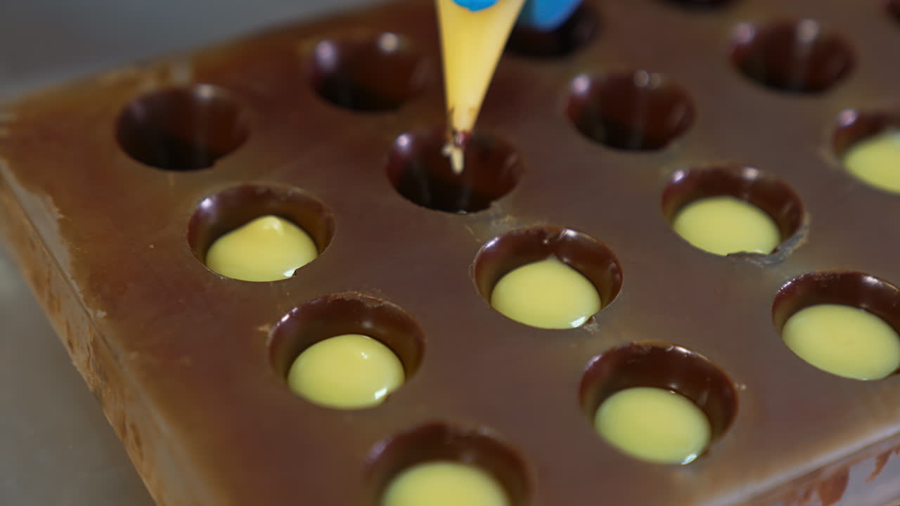 Molds already covered with chocolate are filled with white stuffing. Gloved hand squeezes the filling into the molds from pastry bag. Close up.