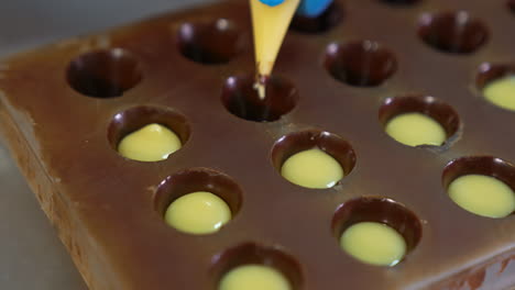 molds already covered with chocolate are filled with white stuffing. gloved hand squeezes the filling into the molds from pastry bag. close up.