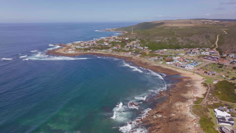 Südafrika-Luftdrohne-Filmischer-Strand-Kleine-Surfstadt-Garden-Route-Still-Bay-Jeffreys-Bay-Häuser-Und-Gebäude-Wellen-Krachen-Auf-Riff-Menschen-Gehen-Am-Späten-Vormittag-Nachmittags-Auf-Der-Straße-Rückwärtsbewegung