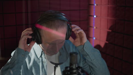 un hombre en un estudio de grabación con una camisa de manga larga azul y auriculares negros, ajustando sus auriculares mientras se prepara para grabar con un fondo de panel acústico rojo, micrófono en primer plano