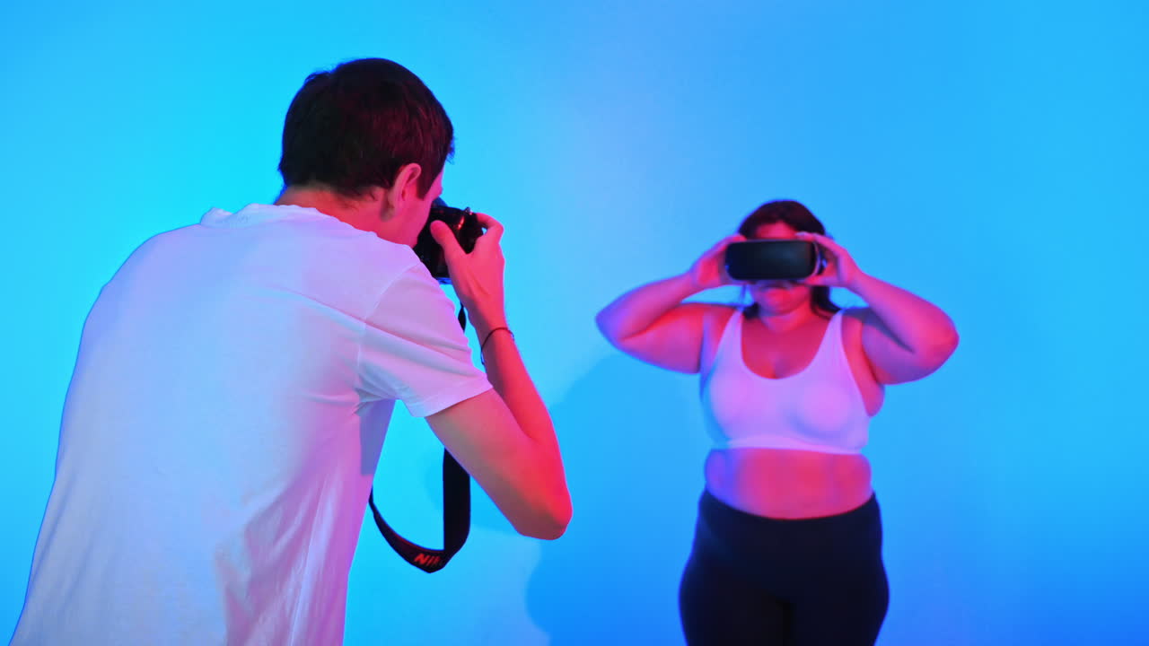 Veduta di un fotografo che scatta ritratti di una donna in sovrappeso con una tuta da ginnastica e un visore VR in uno studio con sfondo blu e illuminazione rossa